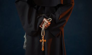 Male monk in black robe holds wooden rosary and cross in hands, religion. Mysterious friar in dark cape
